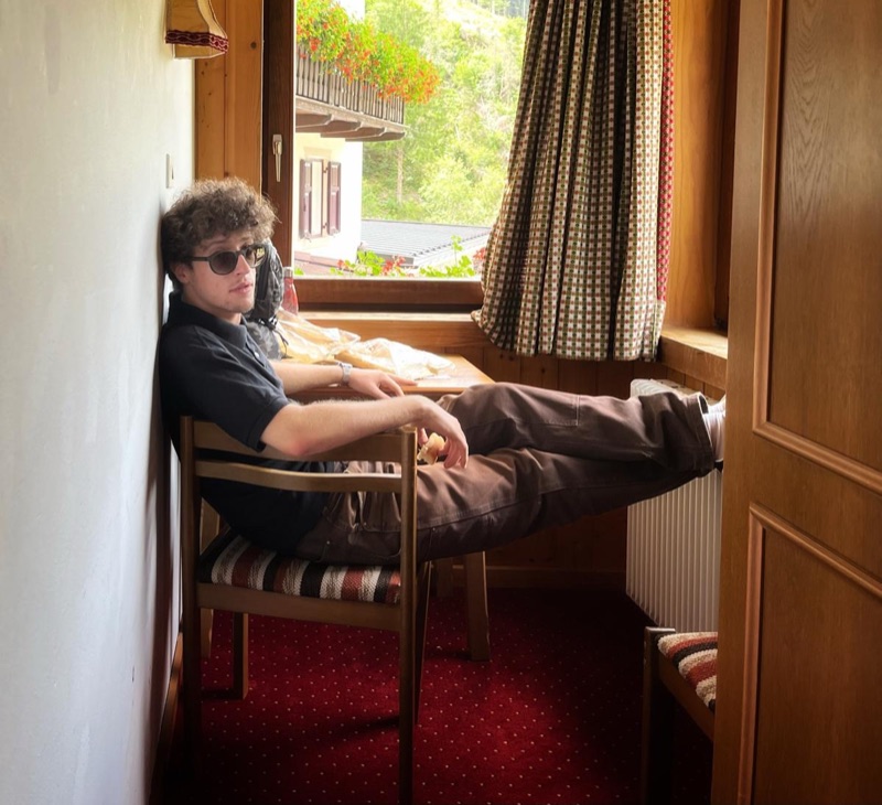 Edoardo with sunglasses, feet up on a chair by the window of a wood-panelled mountain room